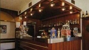 A bar area with wooden paneling, a counter with various bottles, and a ceiling with exposed beams and lights. - Bellavista (Comelico Superiore)