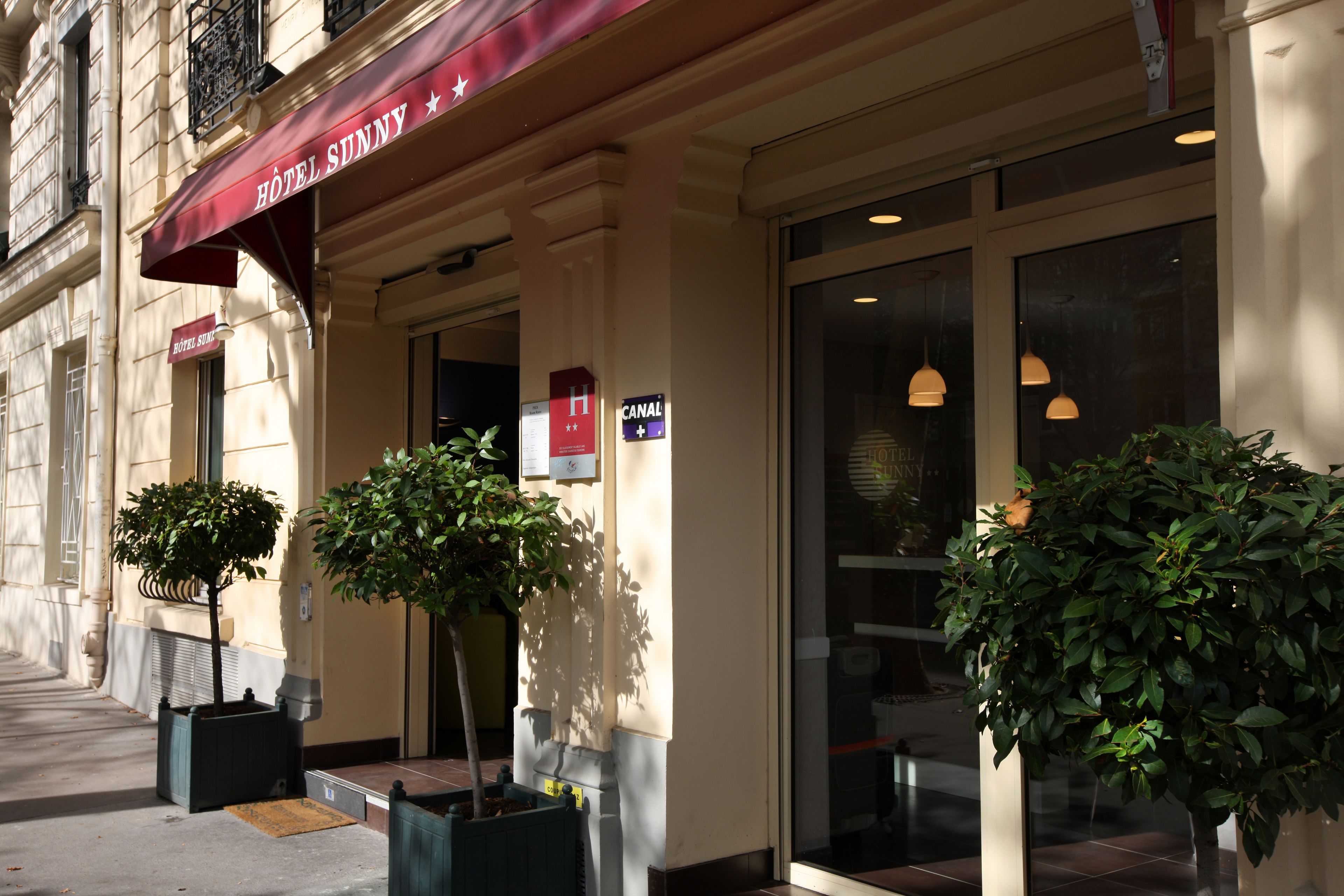 A hotel entrance with a red awning, a sign for 'Hôtel Sunny', and two potted plants.