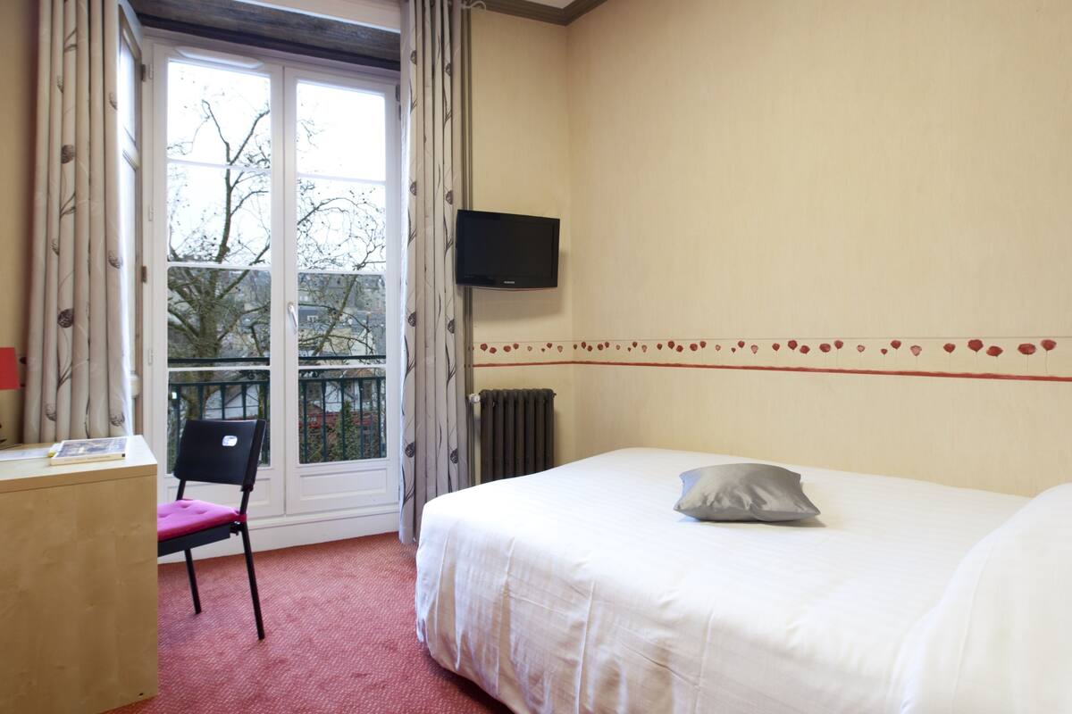 Habitación de hotel con cama, escritorio, silla, televisor y ventana con vistas a árboles y un balcón.