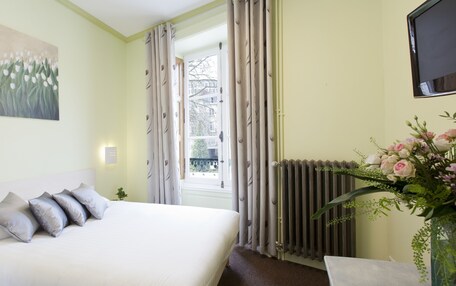 A bedroom with a bed, a painting, a window with curtains, a radiator, and a vase with flowers.