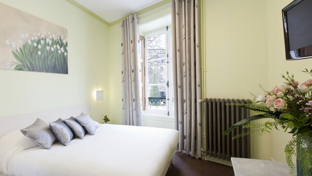 Un dormitorio con cama, un cuadro, una ventana con cortinas, un radiador y un jarrón con flores.