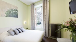 A bedroom with a bed, a painting, a window with curtains, a radiator, and a vase with flowers. - Hôtel Duquesne (Nantes)