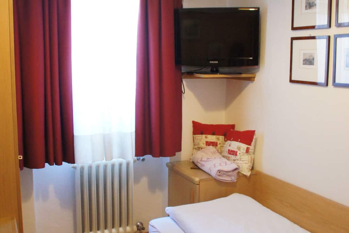 A hotel room with a bed, a television mounted on the wall, a window with curtains, a radiator, and a small table with a chair.