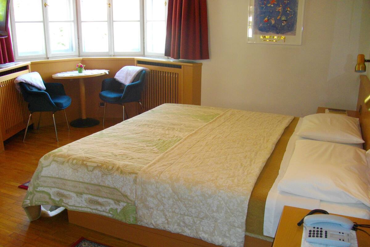 A hotel room with a large bed, a bedside telephone, a small table with two chairs, and a window with curtains.