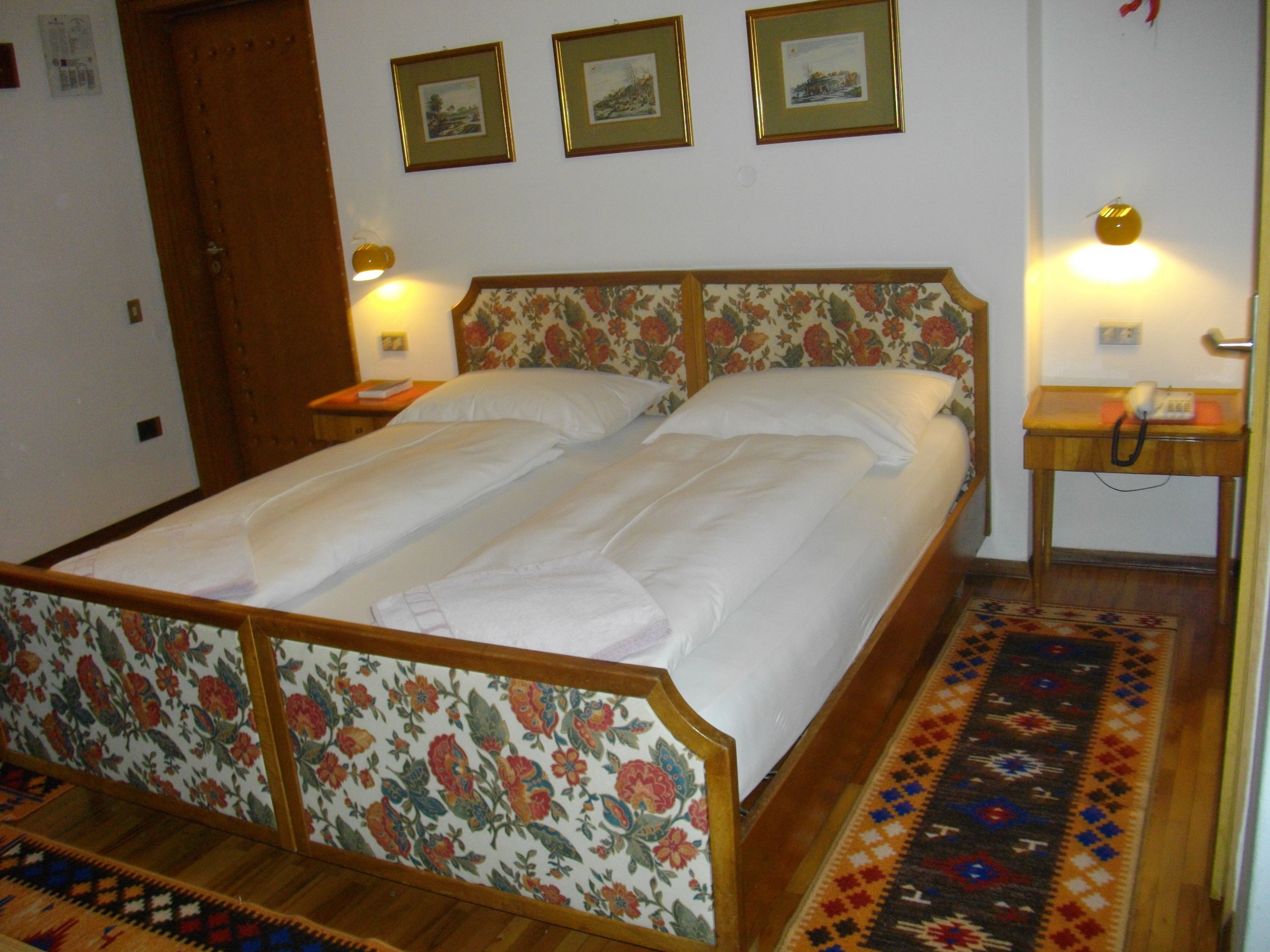 A double bed with floral headboard and footboard, a bedside table with a lamp, and a patterned rug on the floor.