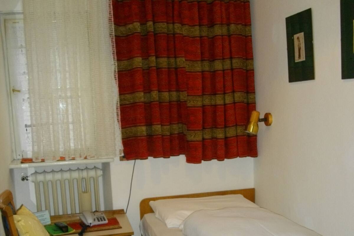 A small, single-bed room with a wooden headboard, a bedside table with a lamp, a chair with a cushion, and a radiator.