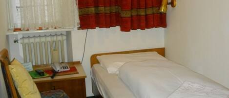 A small, single-bed room with a wooden headboard, a bedside table with a lamp, a chair with a cushion, and a radiator.