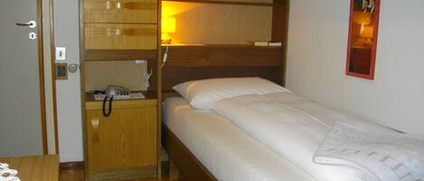 A compact room with a single bed, a bedside table with a lamp, a phone, and a small shelf.