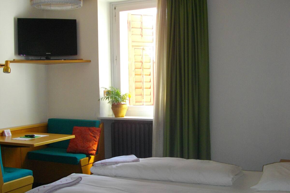 A hotel room with a bed, a desk with a telephone, a television on a shelf, a window with curtains, and a potted plant.