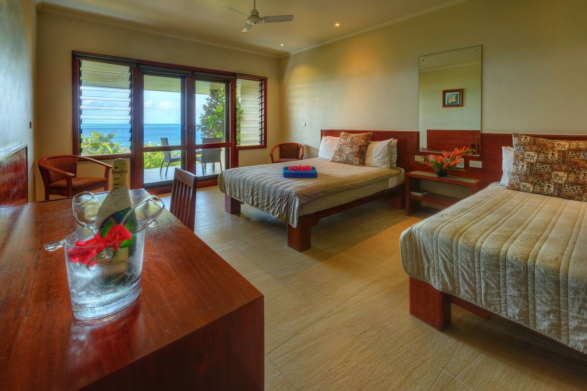 A hotel room with two beds, a desk, and a chair. There is a large window with a view of the ocean.