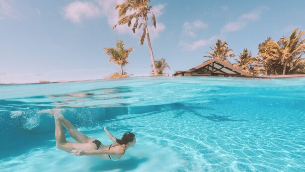 A clear blue pool with a person swimming, surrounded by palm trees and a building with a thatched roof. - Volivoli Beach (Rakiraki)
