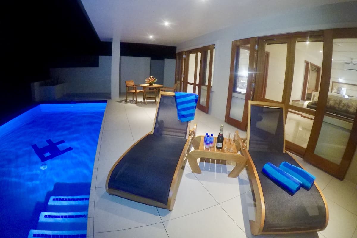 A pool area with lounge chairs and a table, a dining area with chairs and a table, and a kitchen visible through glass doors.