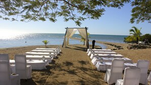 A beach wedding setup with white tables and chairs, a canopy, and a clear view of the ocean.
