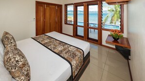 A bedroom with a bed, a wooden wardrobe, a sliding glass door leading to a balcony with a view of the sea and palm trees.