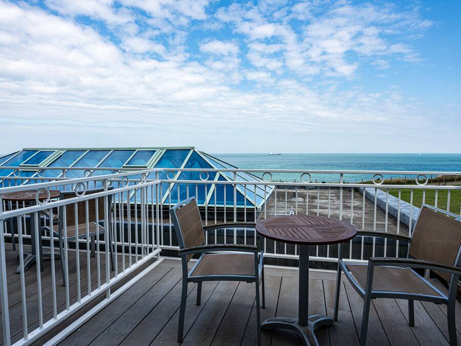 Eine Terrasse mit Tisch und Stühlen, mit Blick auf ein Gewässer und ein Schiff in der Ferne.