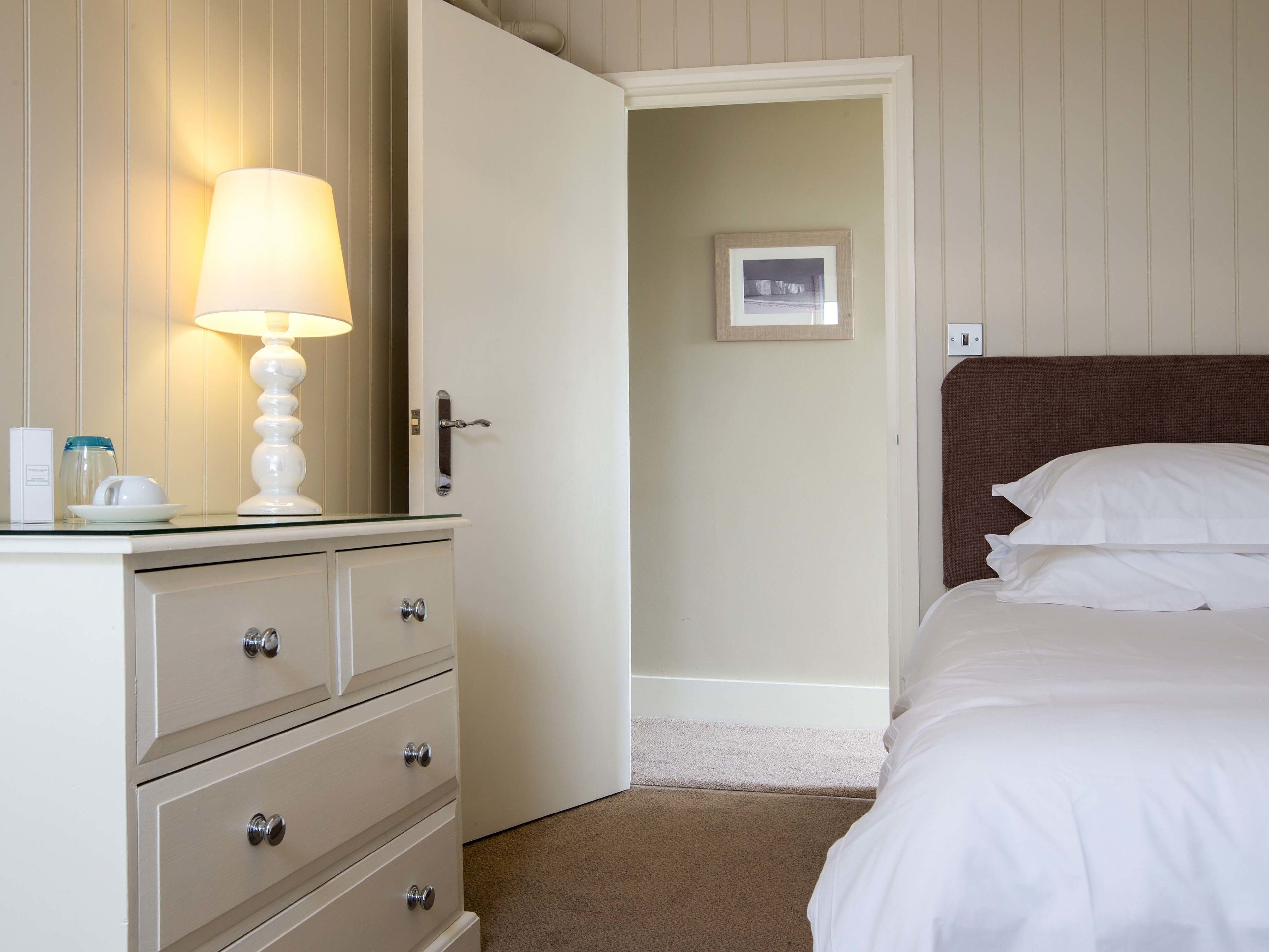 A hotel room with a bed, a bedside table with a lamp, a dresser, and a framed picture on the wall.