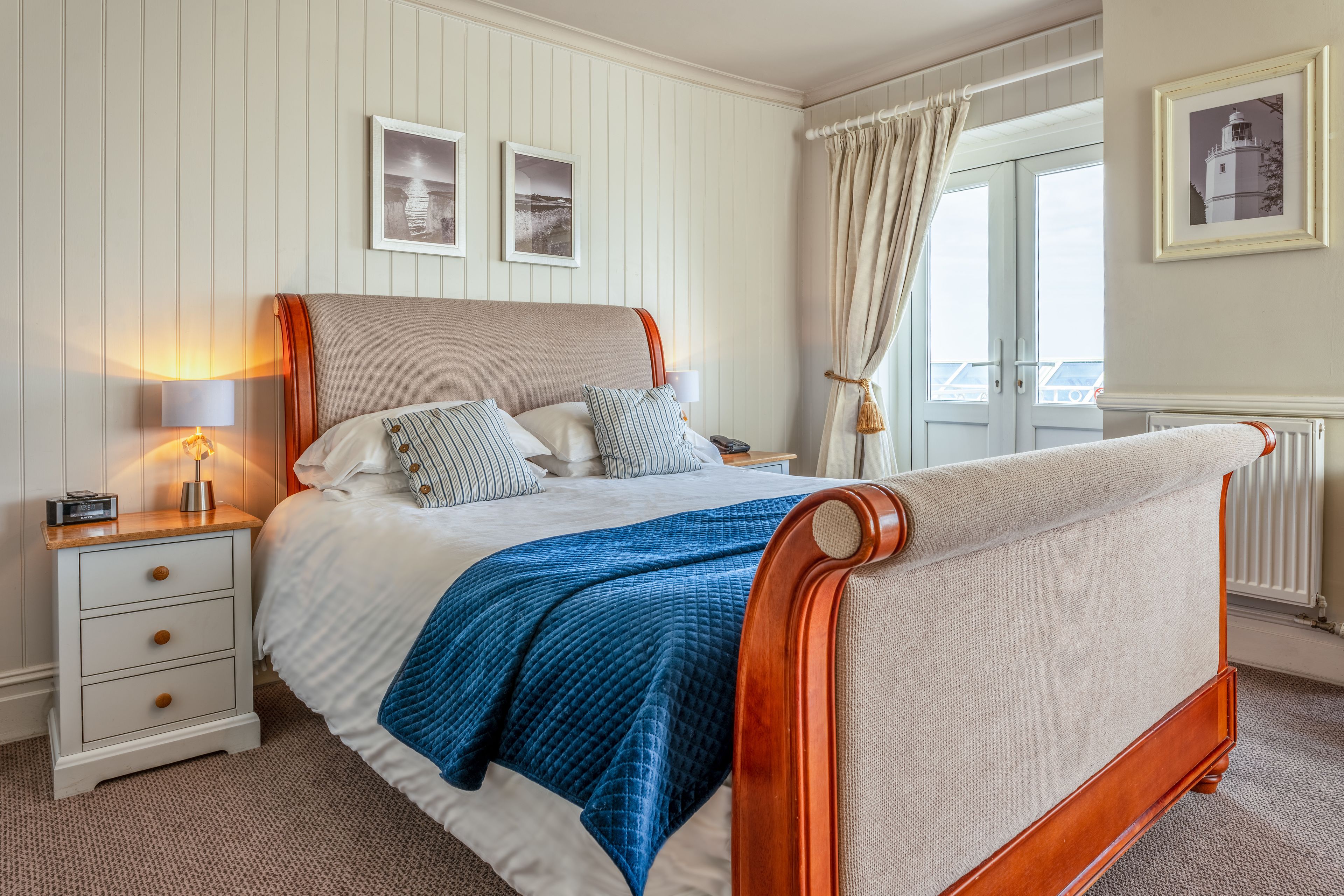 A neatly made bed with a blue quilt, a wooden nightstand with a lamp, and a window with curtains.