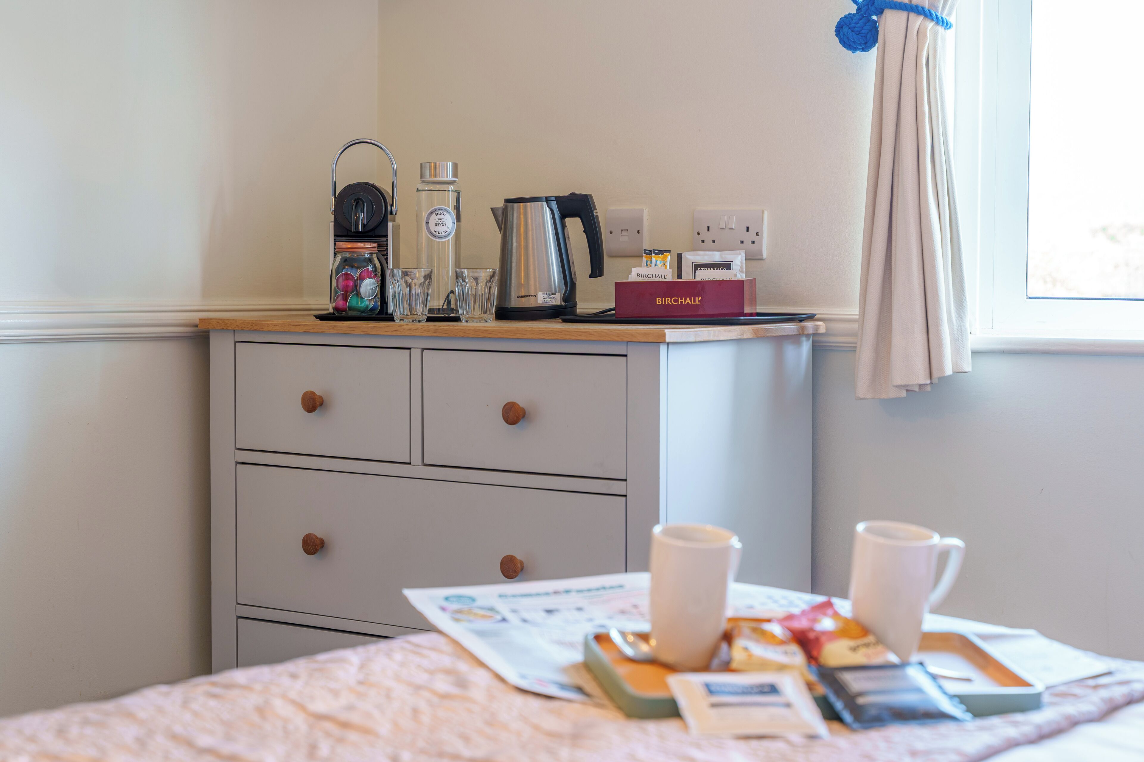 A neatly organized hotel room with a wooden counter, a kettle, a coffee maker, and a Birchall box.