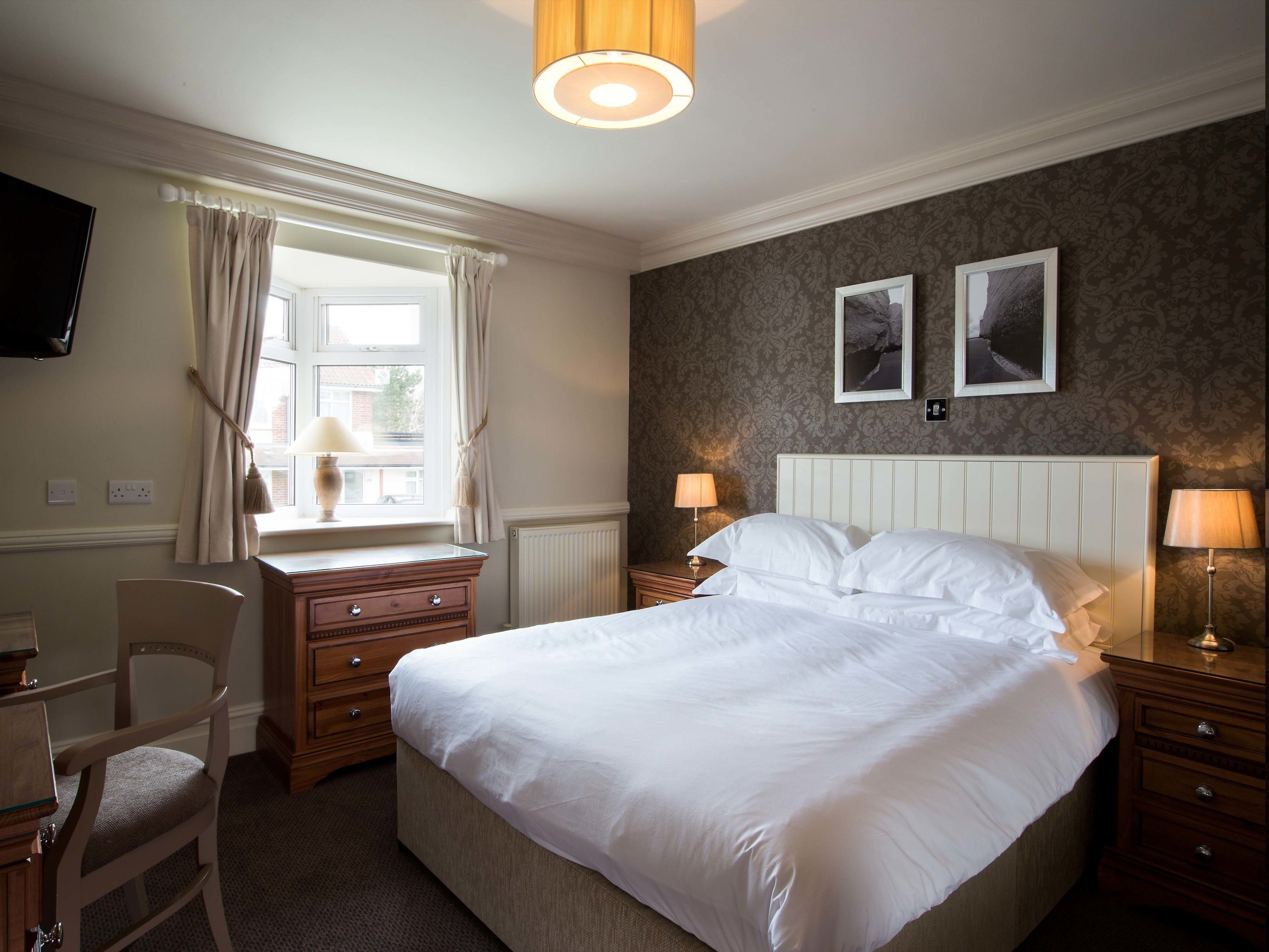 A hotel room with a large bed, two bedside tables with lamps, a chair, a desk, a television, and two framed pictures on the wall.