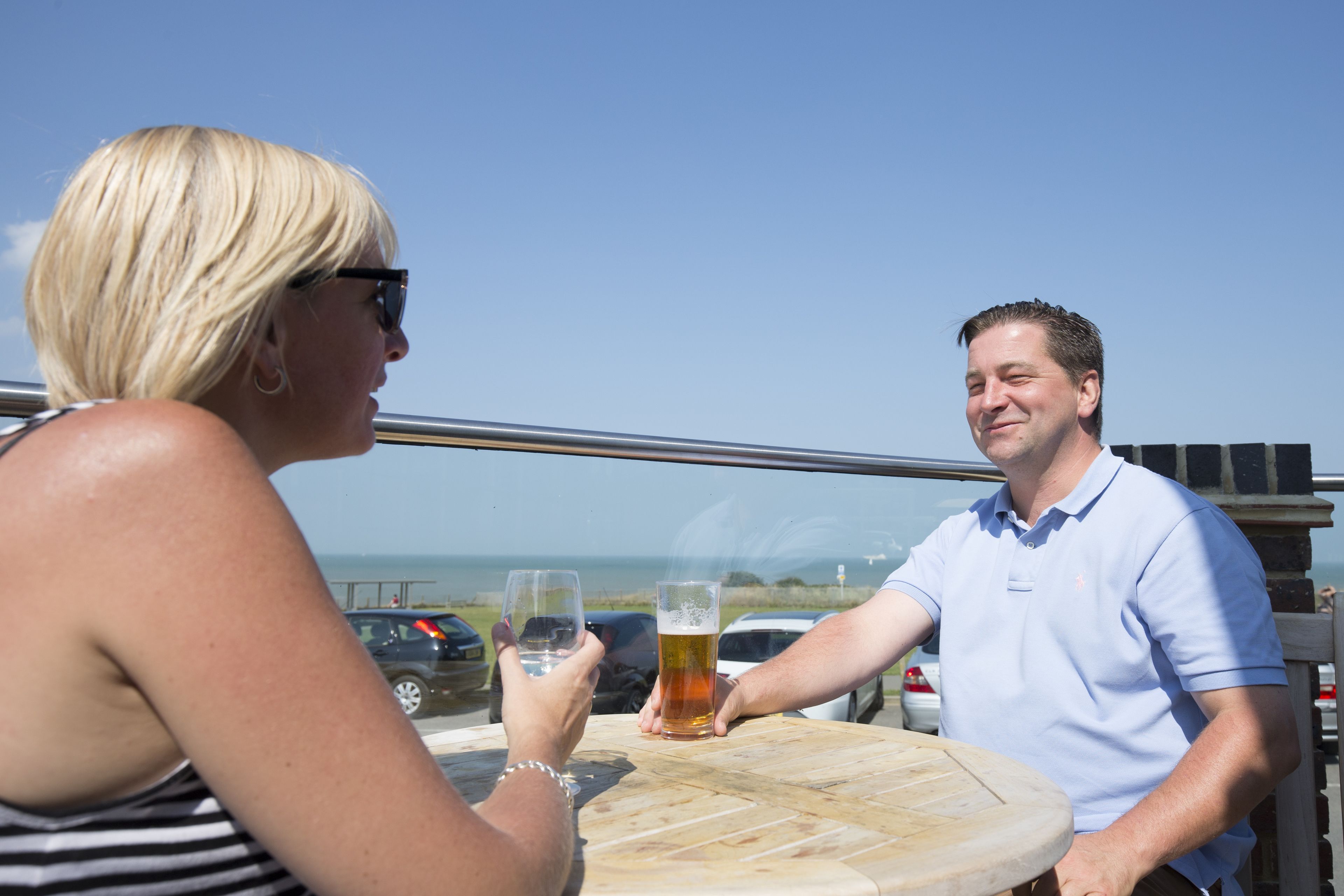A man and a woman toasting with beer glasses at a wooden table with a view of the ocean and parked cars in the background.