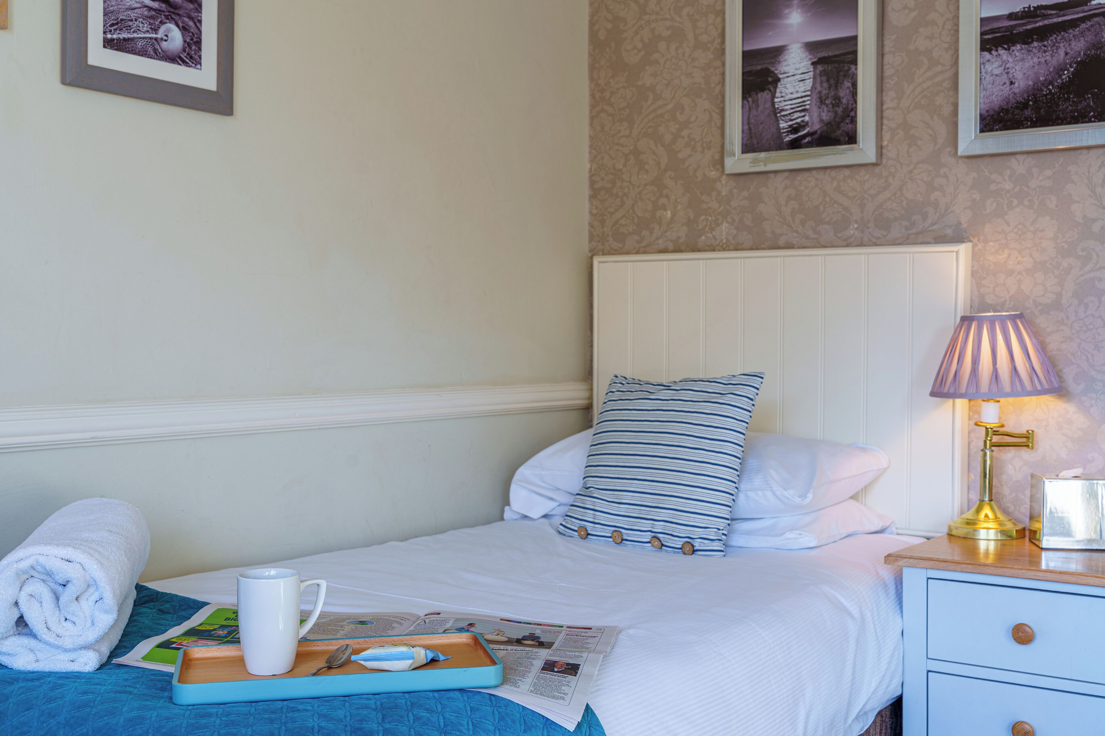 A neatly made bed with a striped pillow, a bedside lamp, and a rolled towel on the floor.