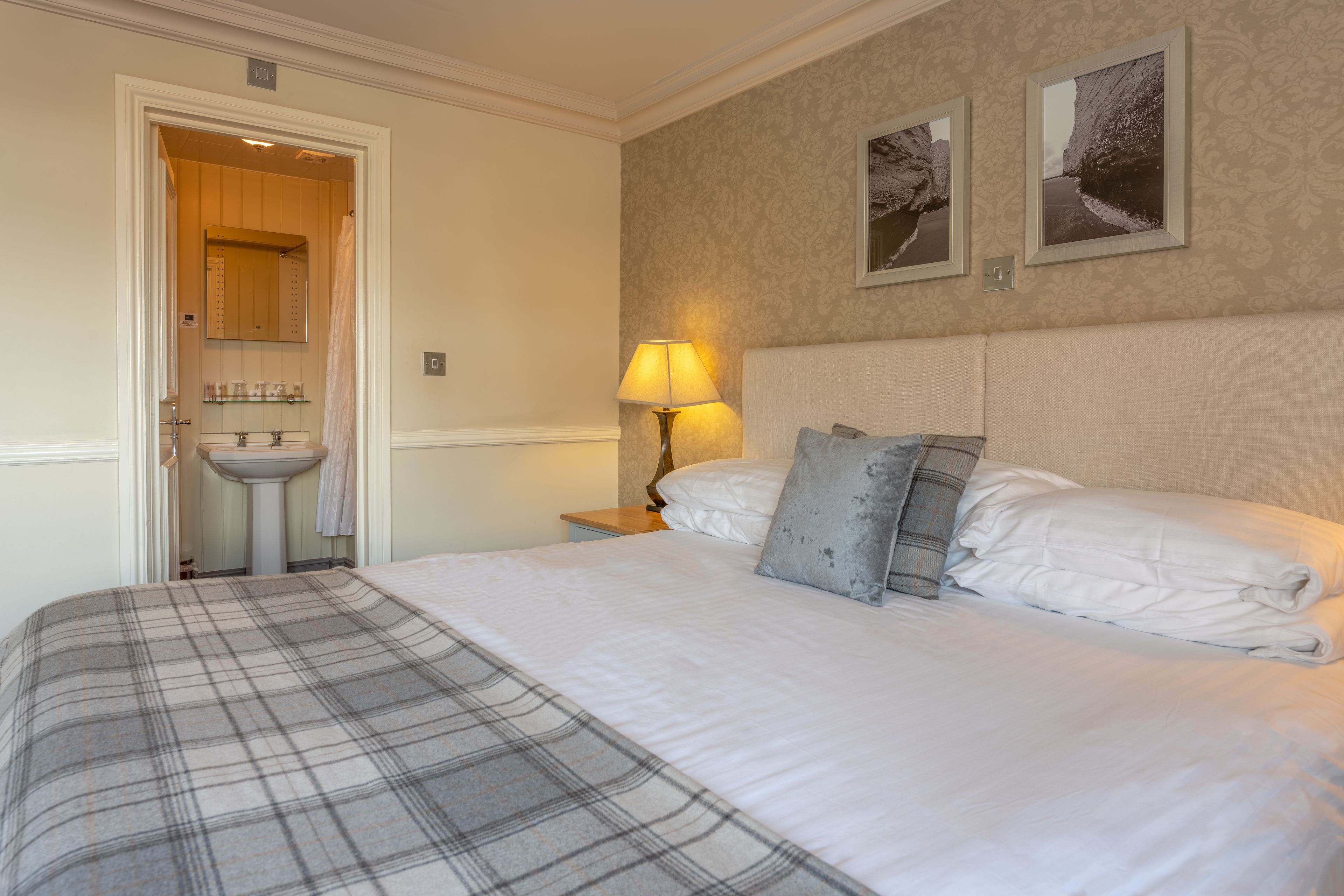 A hotel room with a bed, a nightstand, a lamp, a bathroom with a sink, and two framed pictures on the wall.