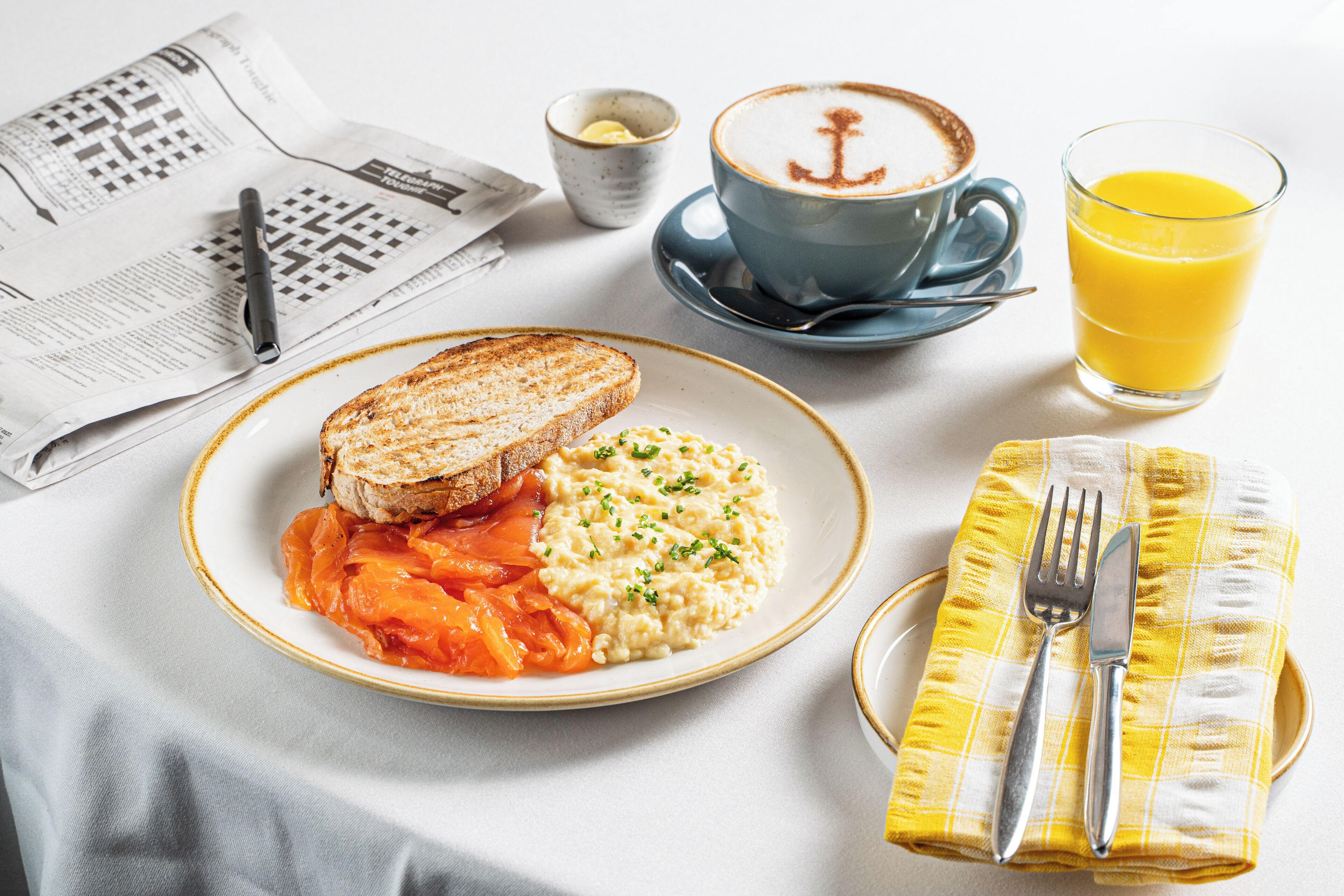 Une table de petit-déjeuner avec des toasts, des œufs brouillés et une tasse de café ornée d’un motif d’ancre, ainsi qu’un journal et un verre de jus d’orange.