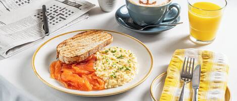 Une table de petit-déjeuner avec des toasts, des œufs brouillés et une tasse de café ornée d’un motif d’ancre, ainsi qu’un journal et un verre de jus d’orange.