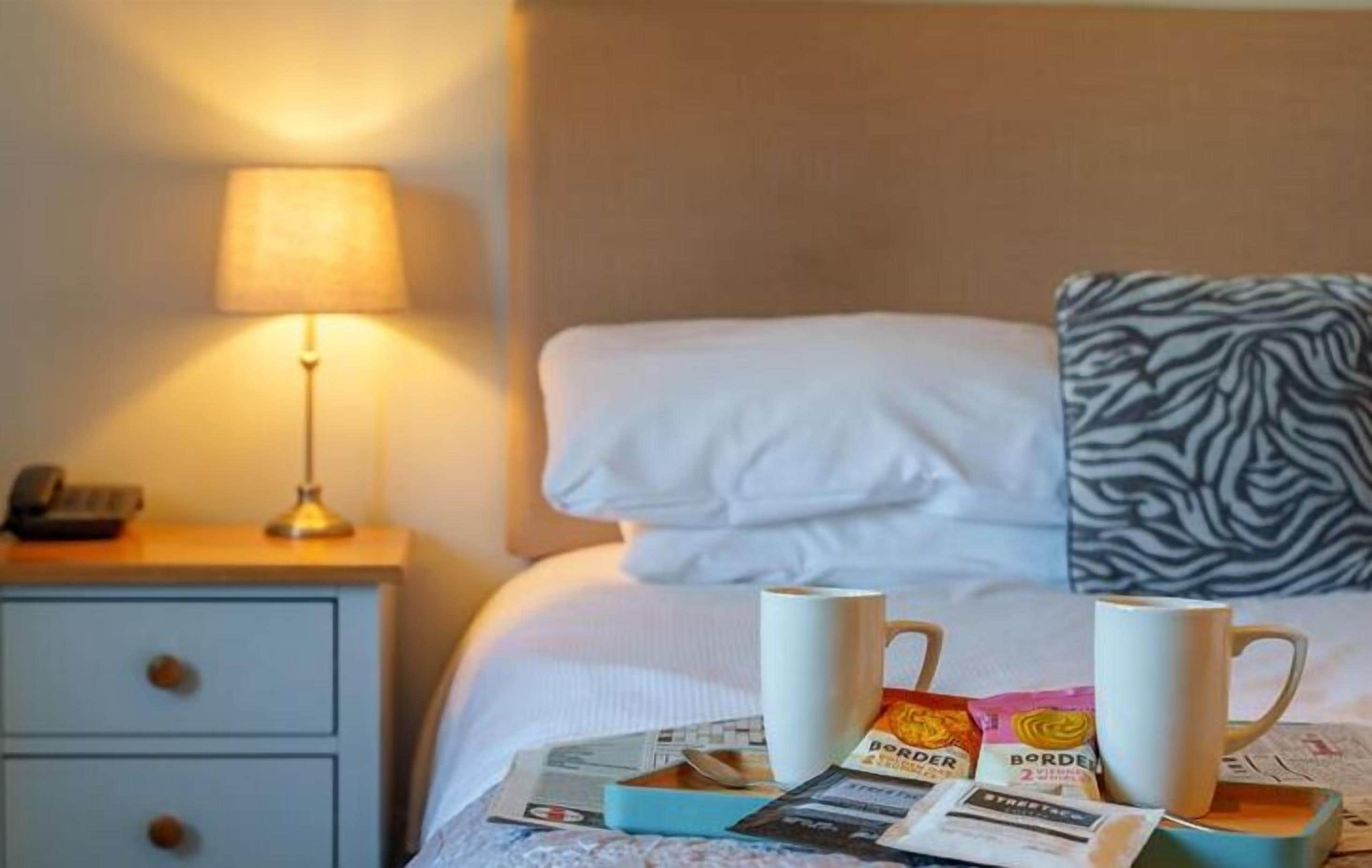 Une chambre d’hôtel avec un lit, une table de chevet avec une lampe et un plateau avec des tasses à café et des éléments pour le petit-déjeuner.
