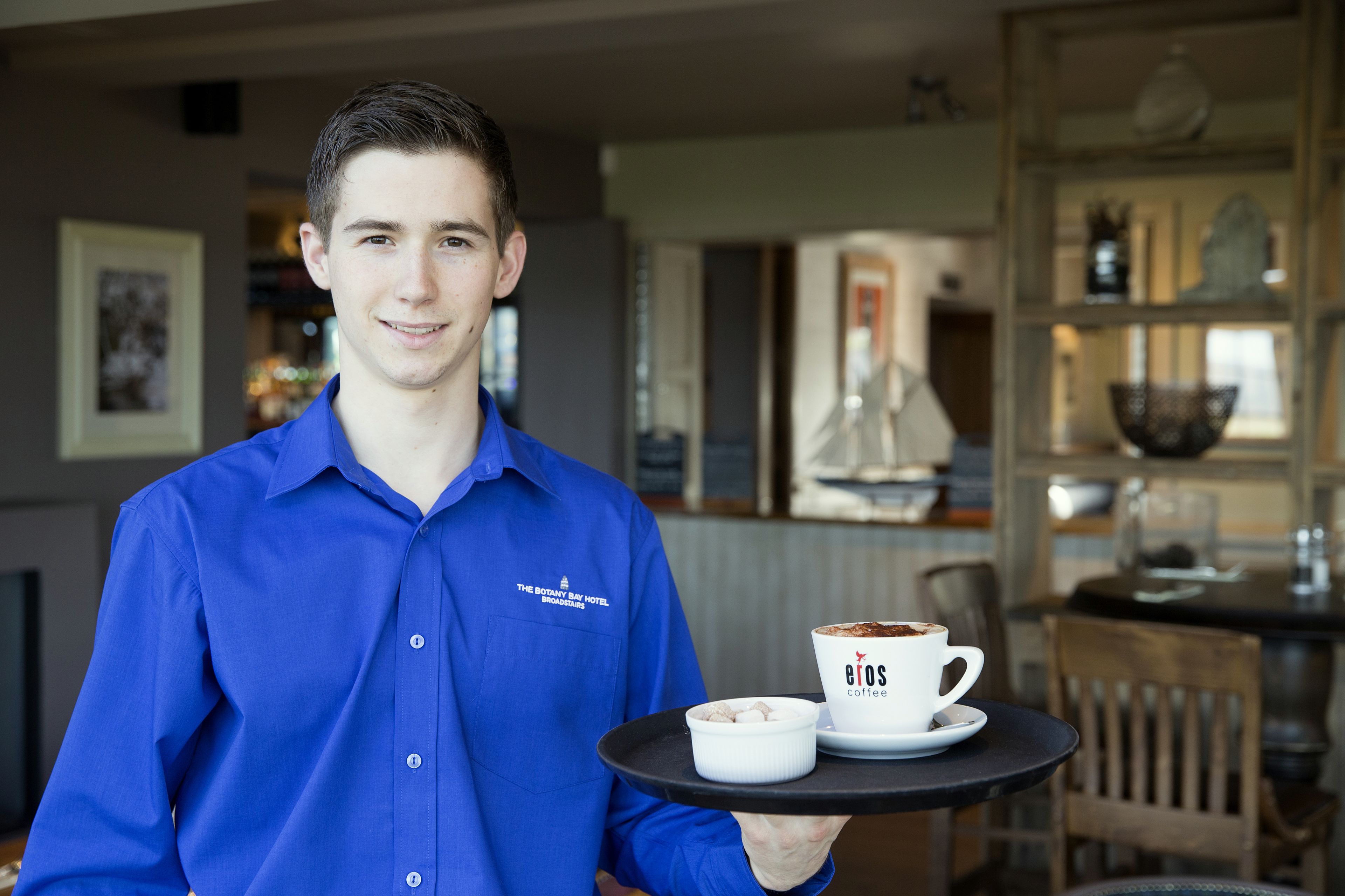 A person in a blue shirt holding a tray with a cup of coffee and a small bowl.