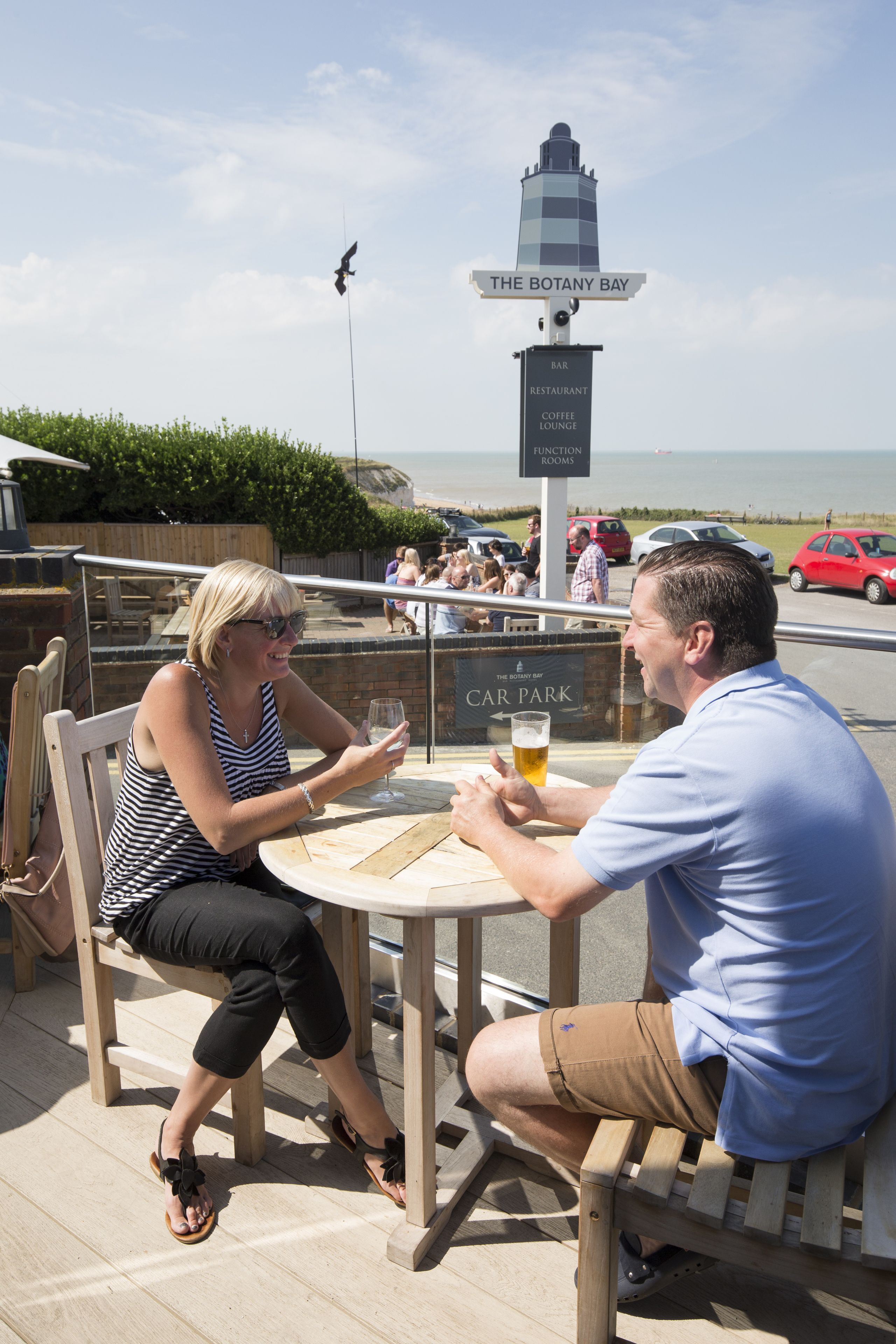 Dos personas sentadas en una mesa con vistas al mar y un cartel que indica 'The Botany Bay'.