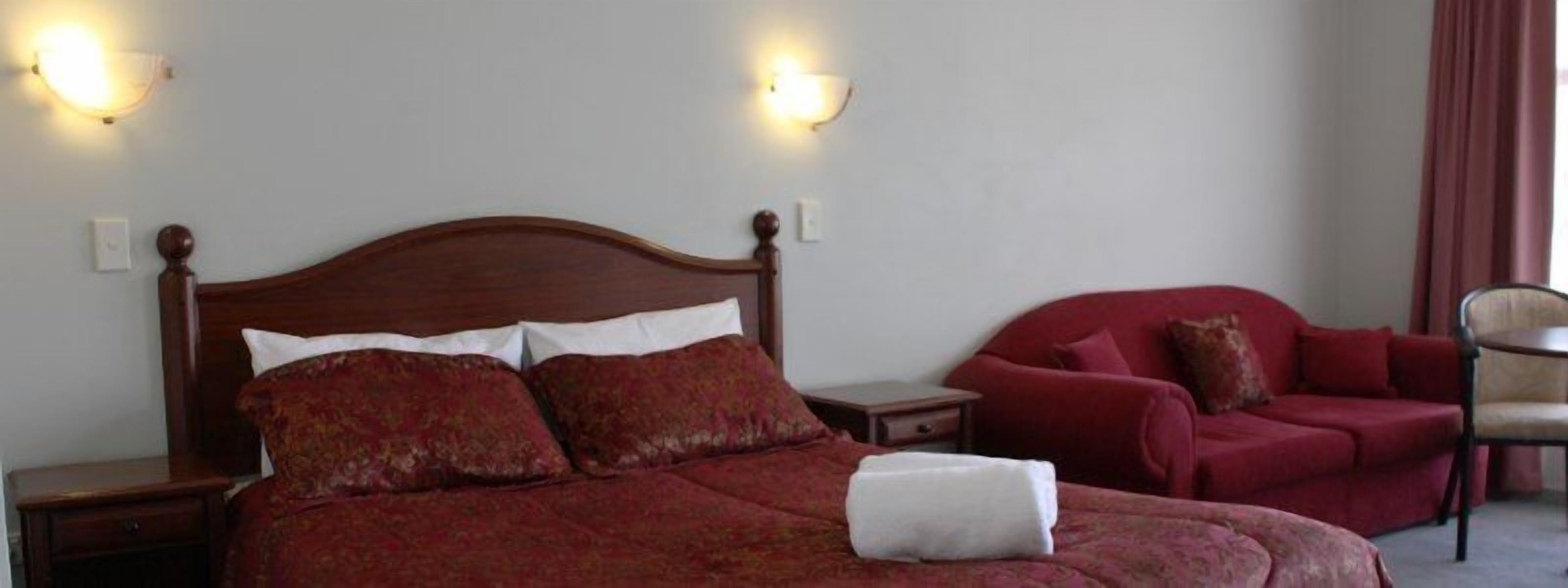 Hotellihuone, jossa on puinen sänky, punaisella kankaalla verhoiltu nojatuoli ja pieni pöytä.