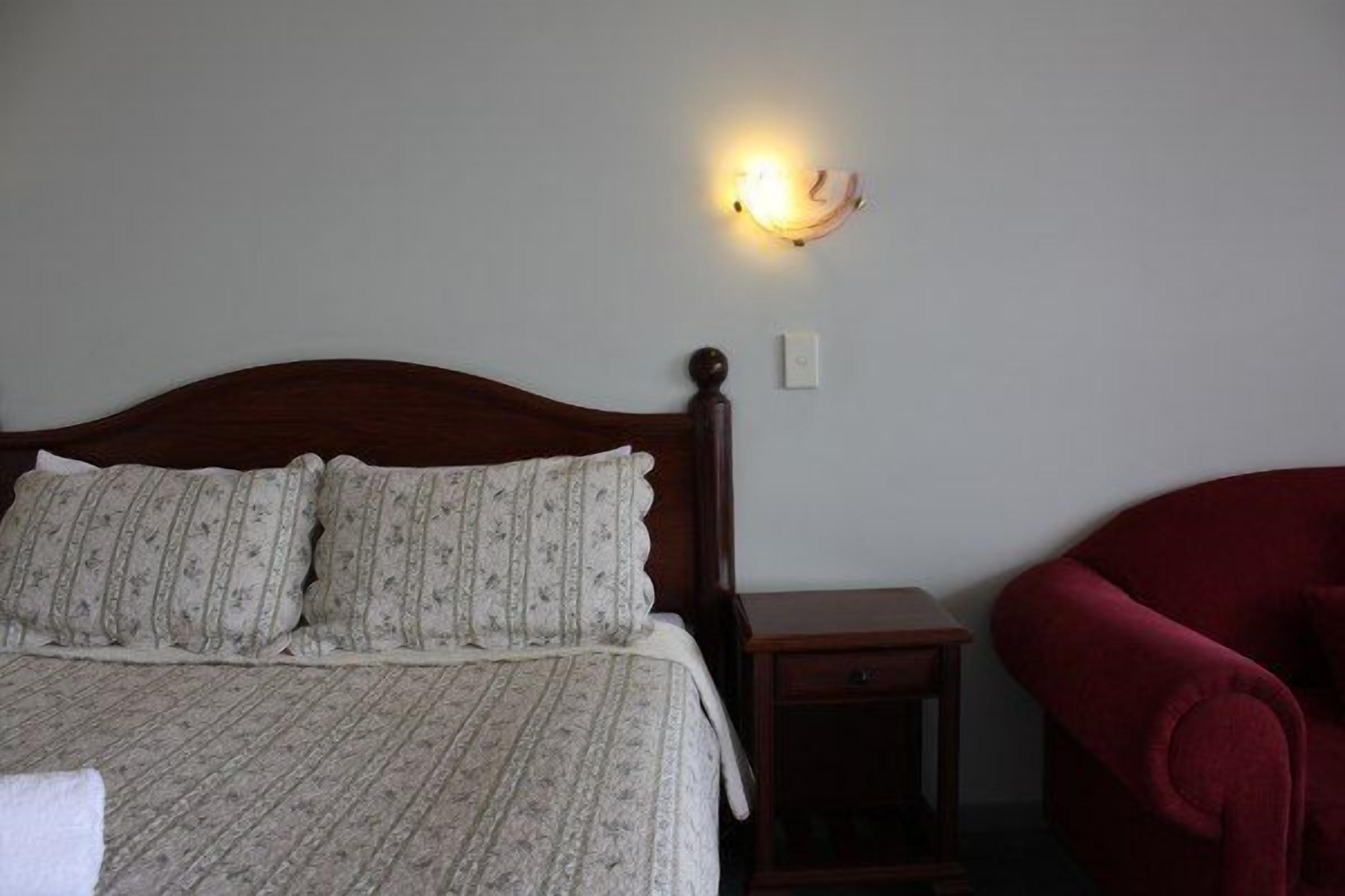 A bedroom with a wooden bed, a nightstand, a red chair, and a wall-mounted light fixture.