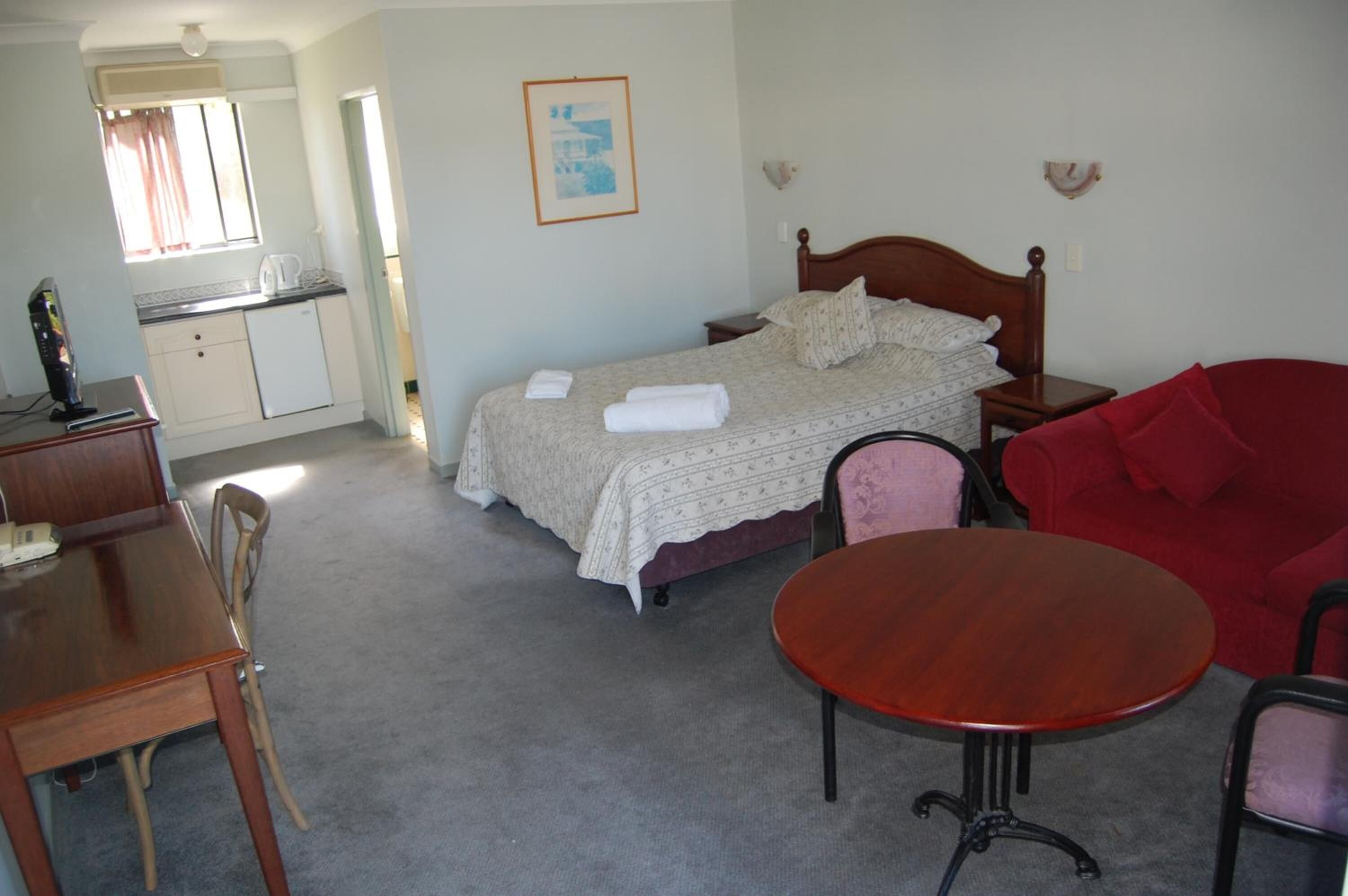 Ein Schlafzimmer mit Bett, Nachttisch, einem kleinen Tisch, einem roten Sofa, einem Holzbüro mit Stühlen und einer Küchenzeile.