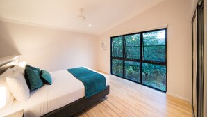 A modern bedroom with a large window, a bed with white linens and teal pillows, and a wooden floor.