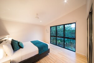 Un dormitorio moderno con un ventanal grande, una cama con ropa de cama blanca y almohadas color turquesa, y piso de madera.