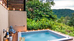A hot tub with candles, a bottle of champagne, and glasses on a wooden deck with a scenic view of a forested hill.