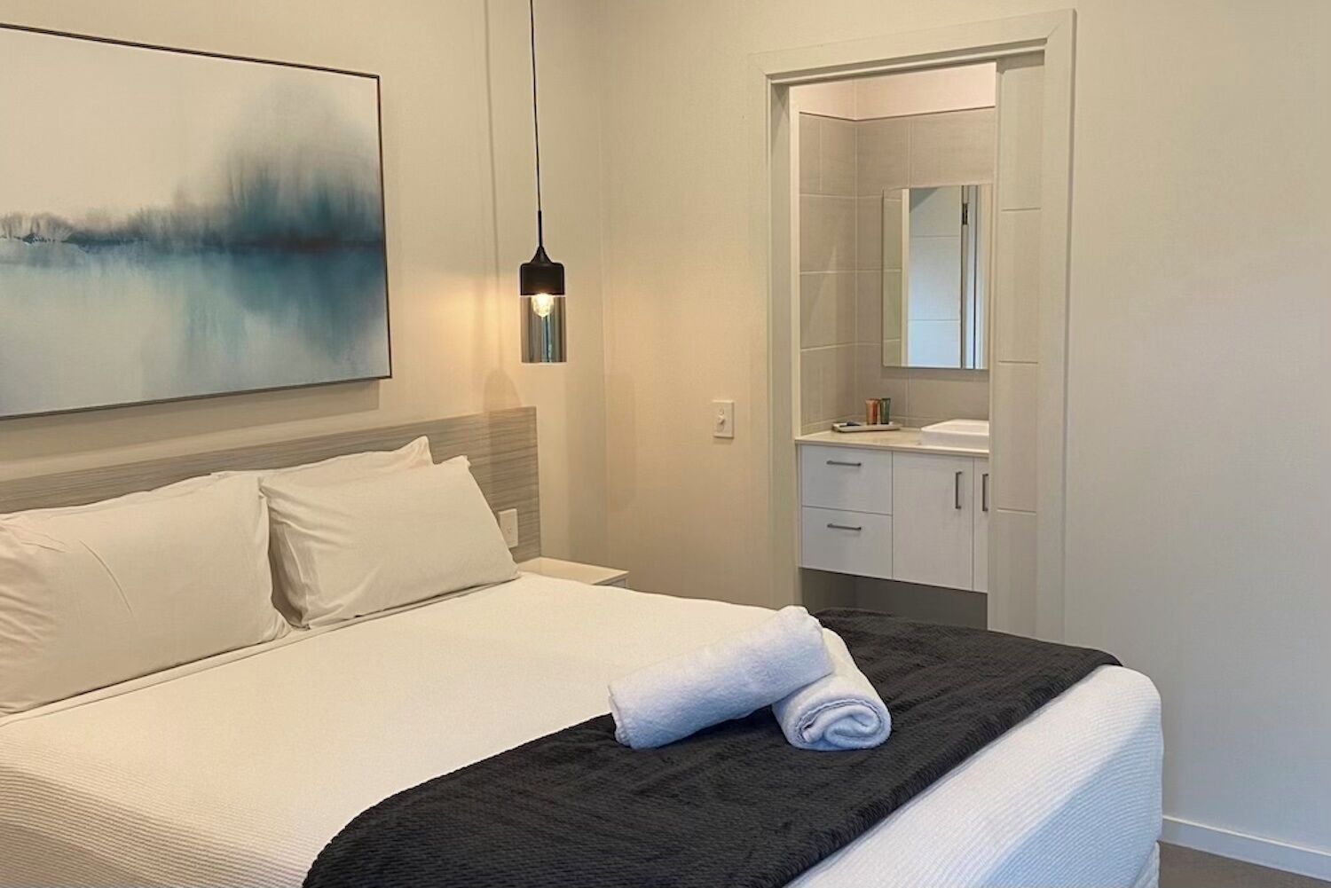 Habitación de hotel con cama, un cuadro, una lámpara colgante y baño con espejo y toalla sobre la cama.