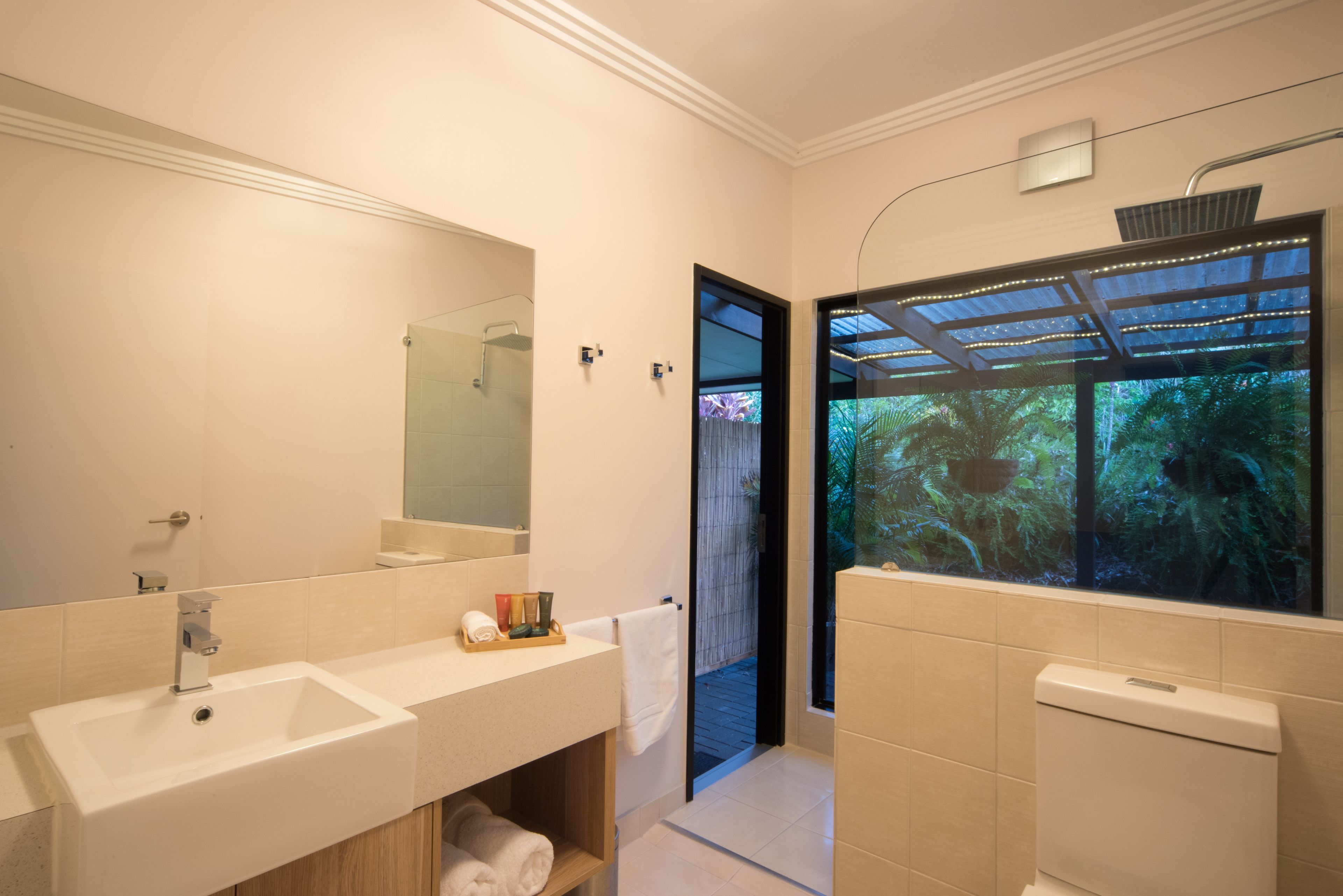 Un baño moderno con un espejo grande, un lavamanos y una ducha. Se ve un ventanal con vistas a la vegetación.