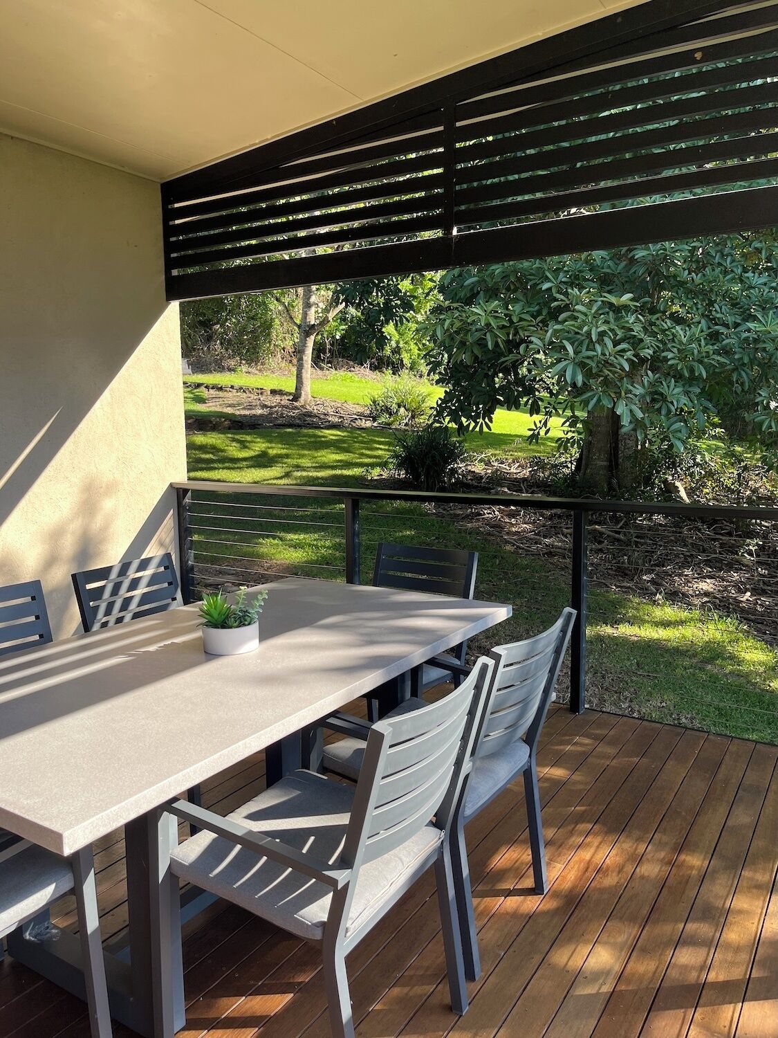 An outdoor patio with a table and chairs, a potted plant, and a view of a garden with trees and grass.