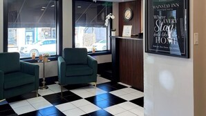 Un hall d’hôtel avec un sol à carreaux, deux fauteuils verts, un comptoir d’accueil, une horloge murale et un panneau encadré.
