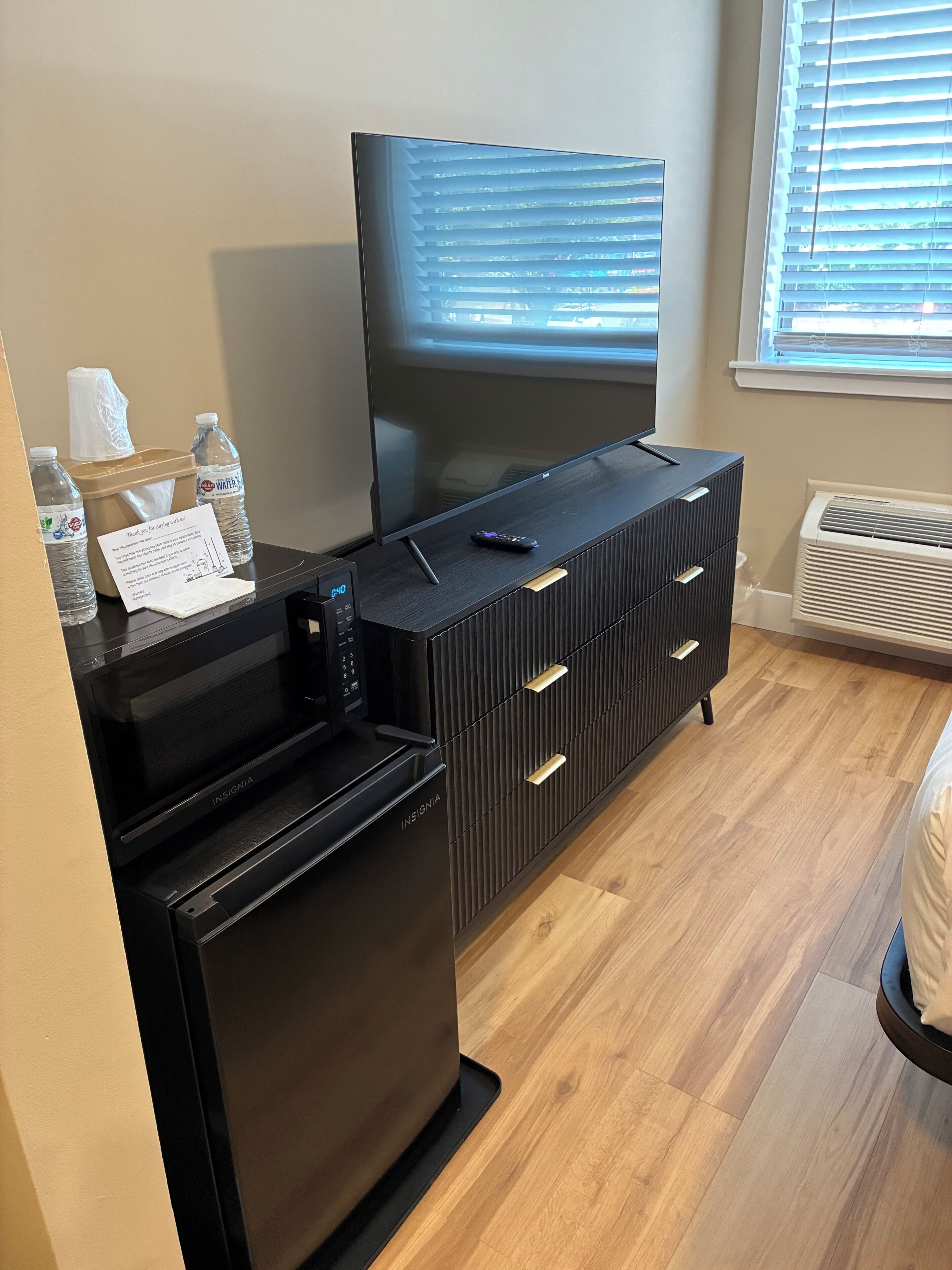 A modern hotel room with a flat-screen TV on a black entertainment unit, a mini-fridge, a remote control, and a note thanking guests for using the air conditioning.