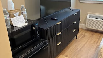 A modern hotel room with a flat-screen TV on a black entertainment unit, a mini-fridge, a remote control, and a note thanking guests for using the air conditioning.