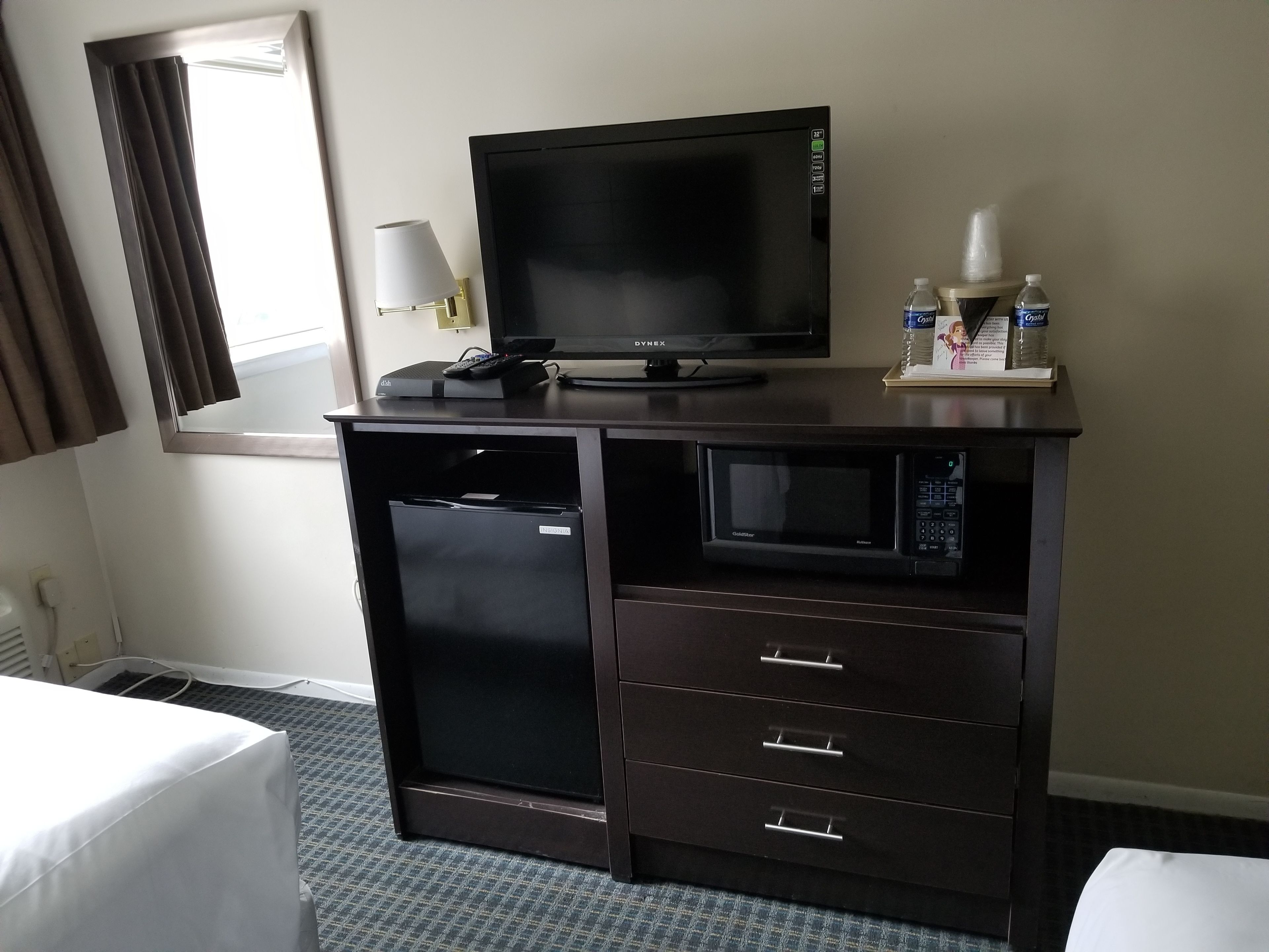 Ein Hotelzimmer mit dunklem Holztisch, Flachbildfernseher, Lampe und Minibar.