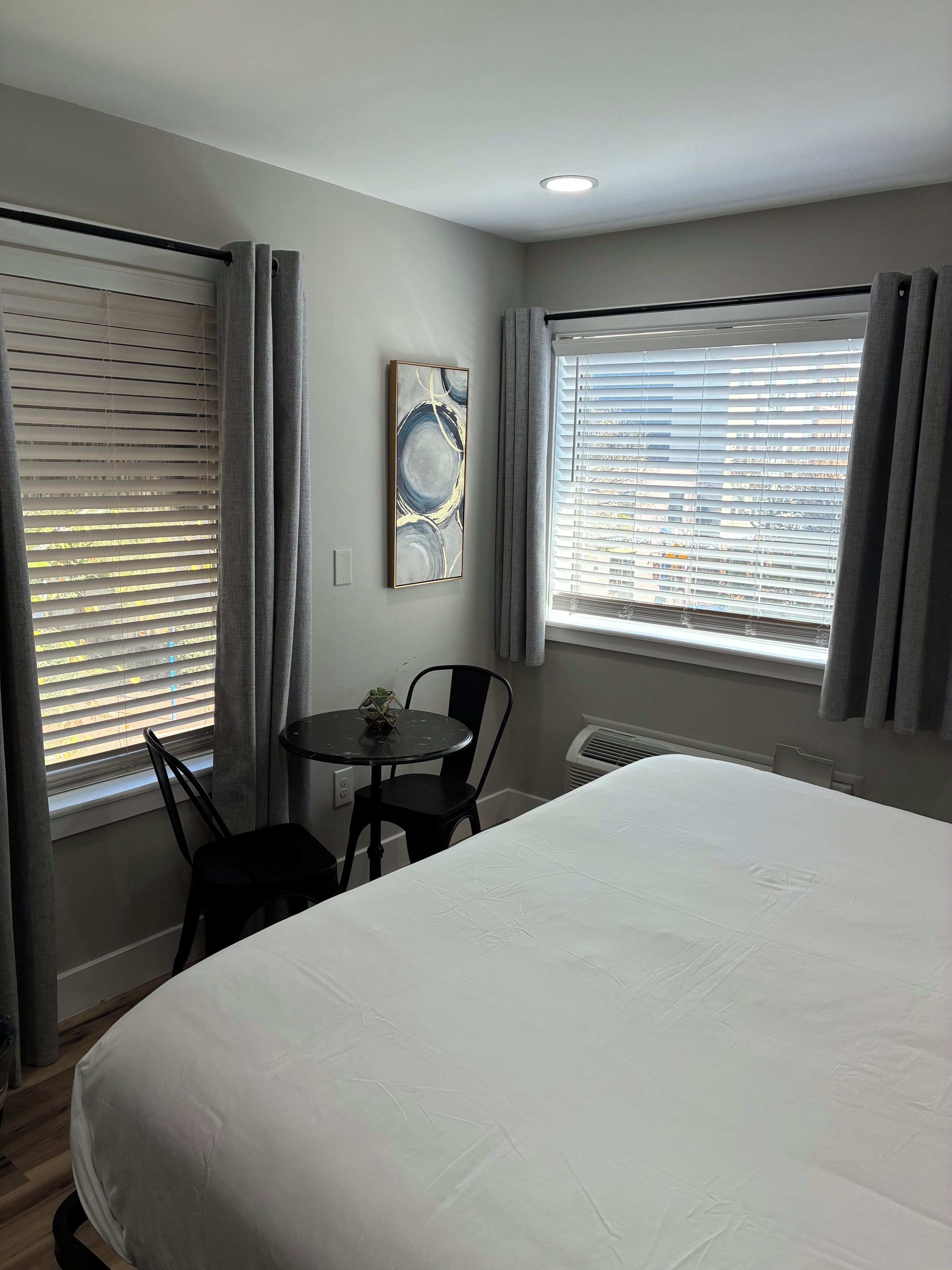 A hotel room with a bed, a small table with a chair, a window with blinds, and a wall-mounted artwork.