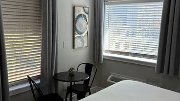 A hotel room with a bed, a small table with a chair, a window with blinds, and a wall-mounted artwork.
