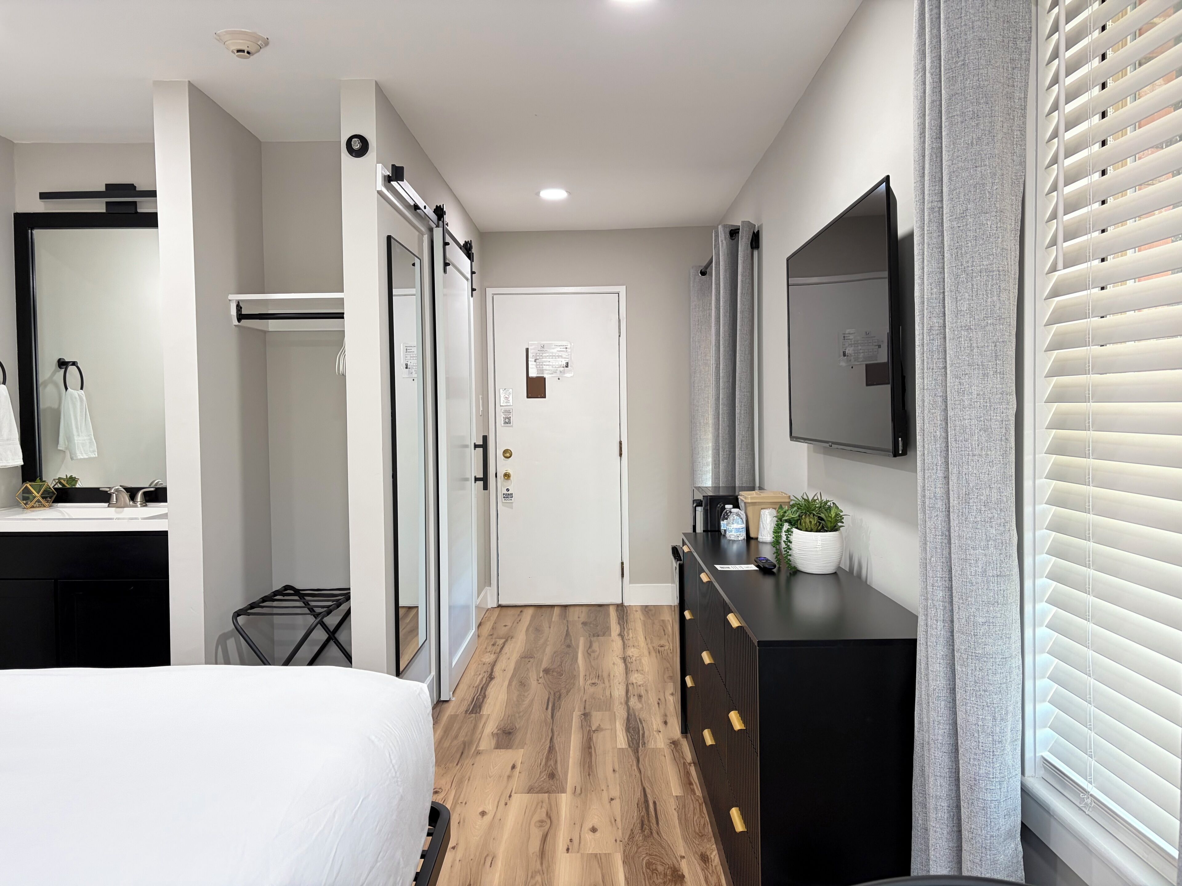 A modern hotel room with a wooden floor, a bed, a black dresser, a mirror, a television, and a door with a towel hanging on it.
