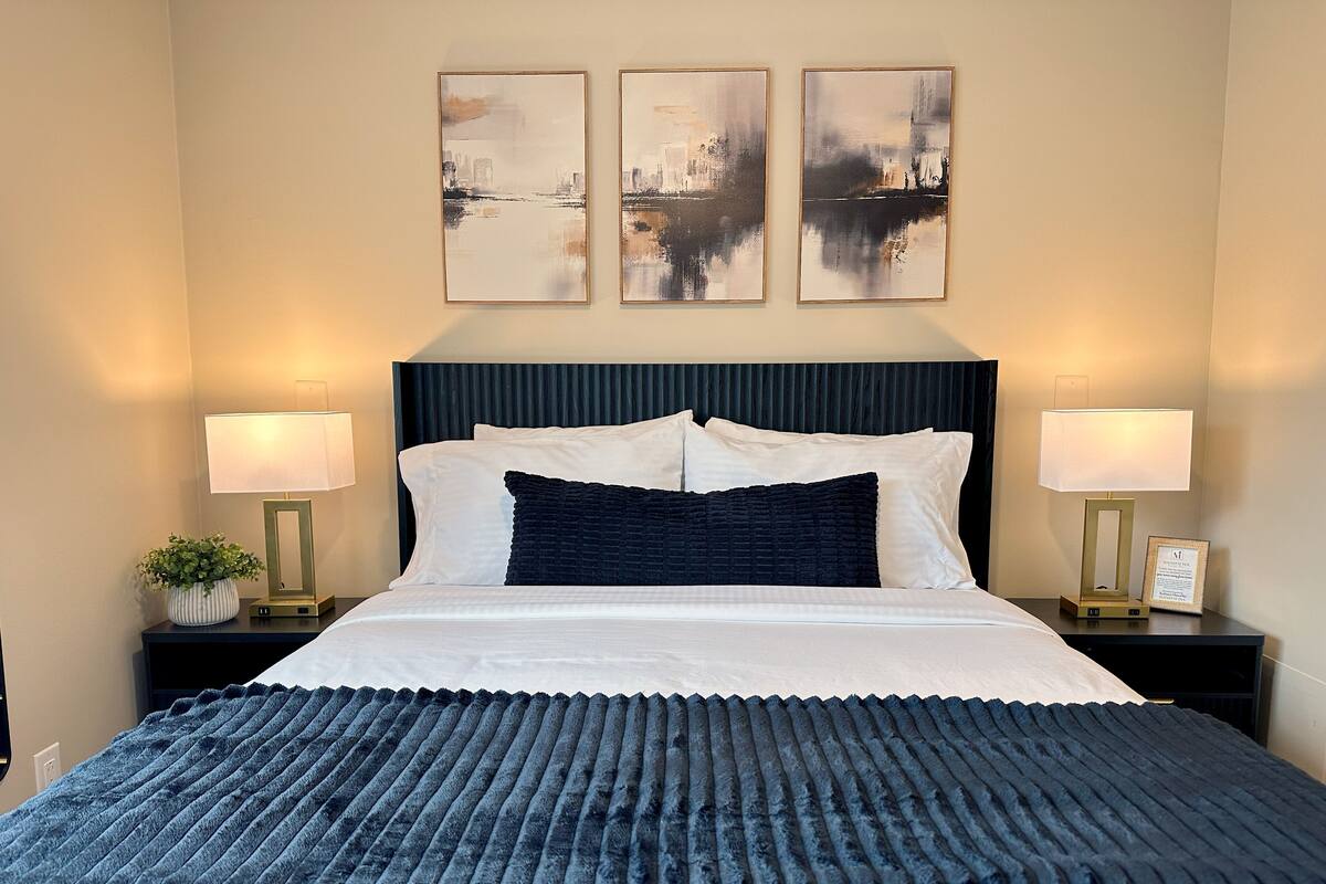 A neatly made bed with a dark blue blanket, two bedside tables with lamps, and three framed artworks on the wall.