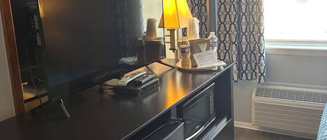A hotel room with a large flat-screen TV, a lamp, a microwave, and a heater near the window.