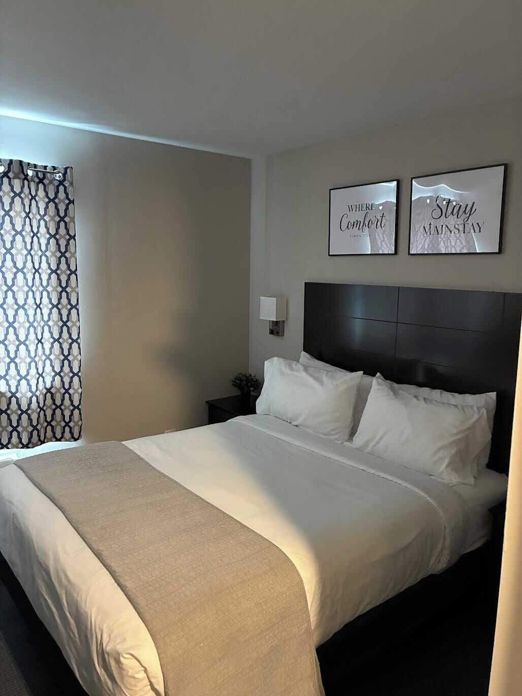 Una habitación de hotel con una cama grande, cabecera, mesita de noche con lámpara y dos citas motivacionales enmarcadas en la pared.